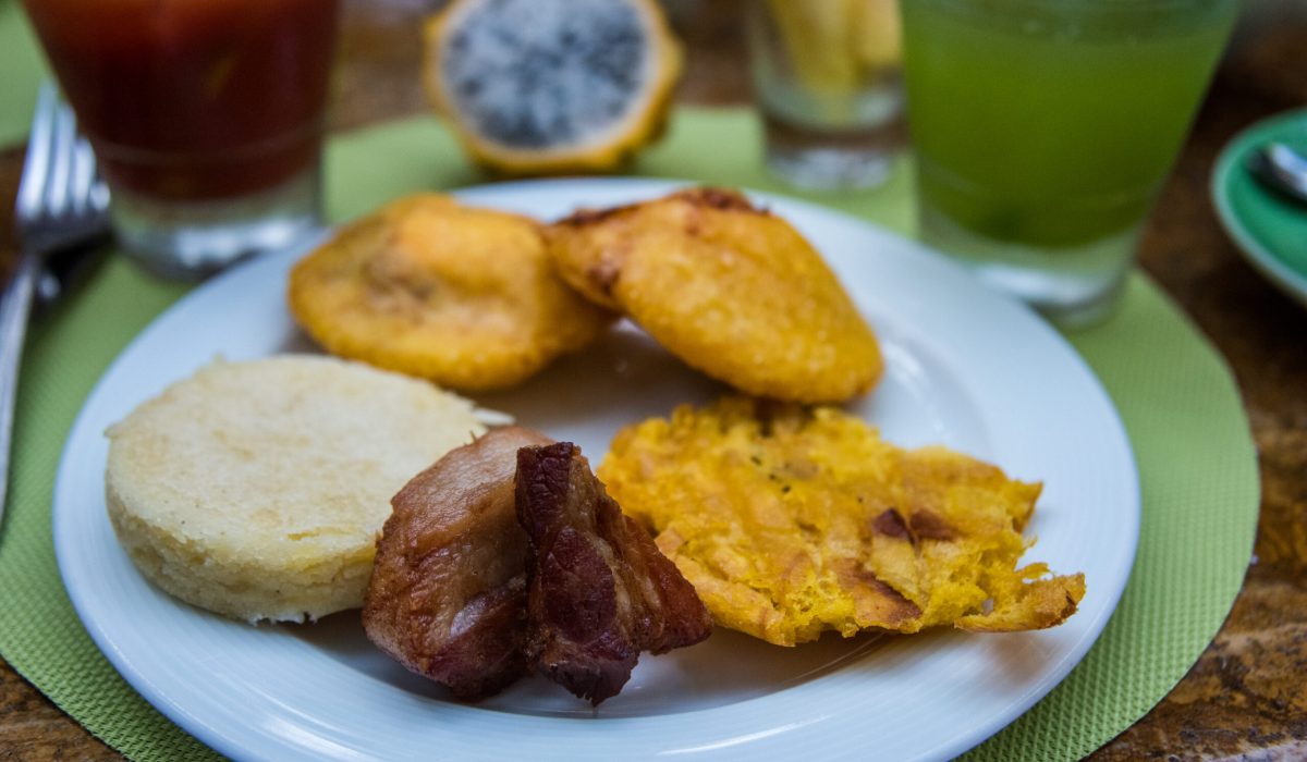 A plate with Colombian breakfast buffet items, arepa, patacones, egg empanada and pitaya and a glass of Bloody Mary cocktail on a table in Cartagena, Colombia