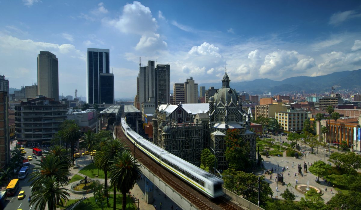 The elevated Medellin Metro is in motion as it glides out of Parque Berrio Metro Station in front of Plaza Botero.  Plaza Botero was named after Medellin's native son and Colombia's most famous artist, Fernando Botero.  The square contains 23 of his most famous sculptures.  The stirped art nouveau Palace of Culture and downtown office buildings can be seen in the background.